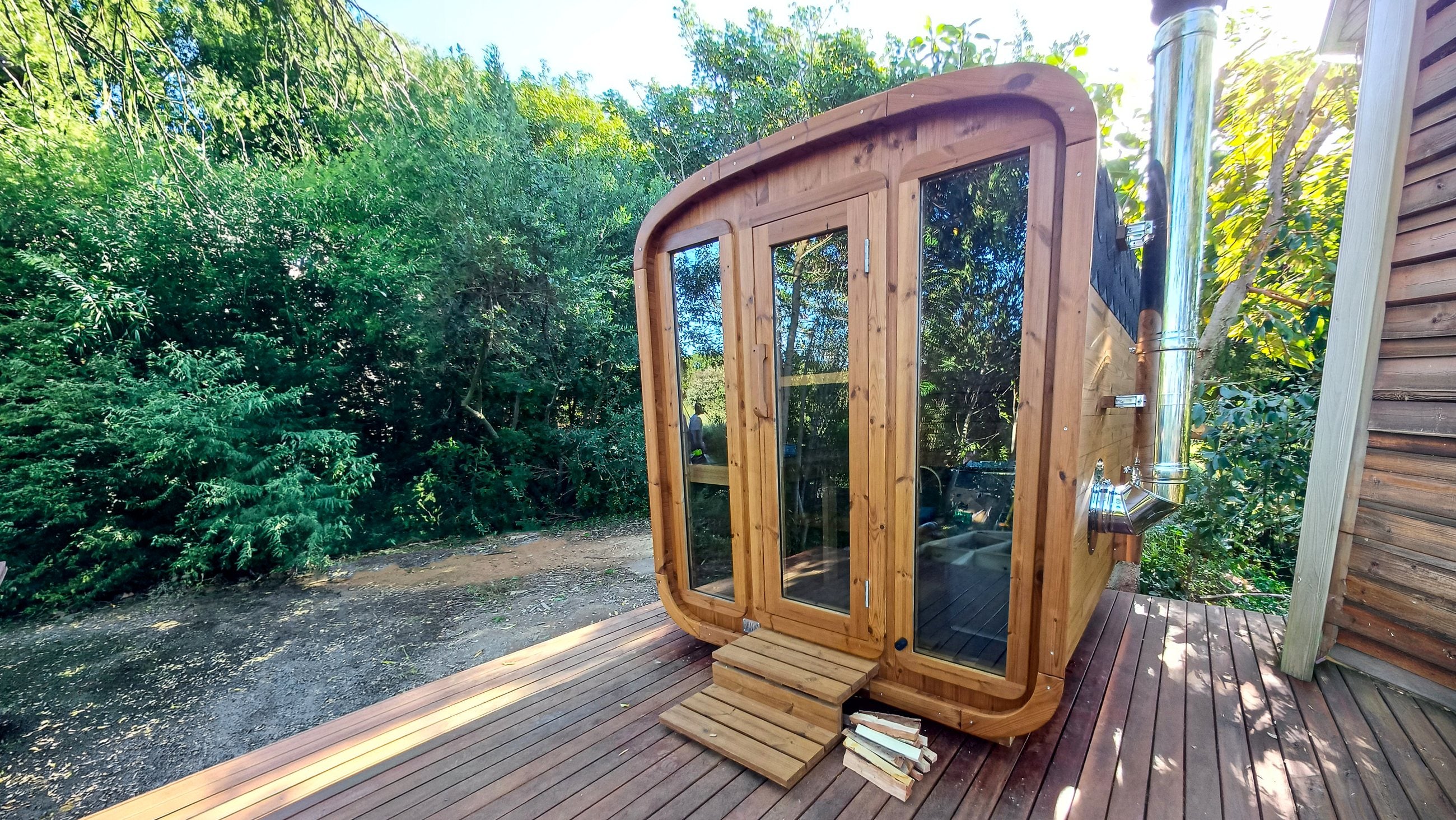 A BANYA Cube on a timber deck, flue fitted and firewood stacked at the door